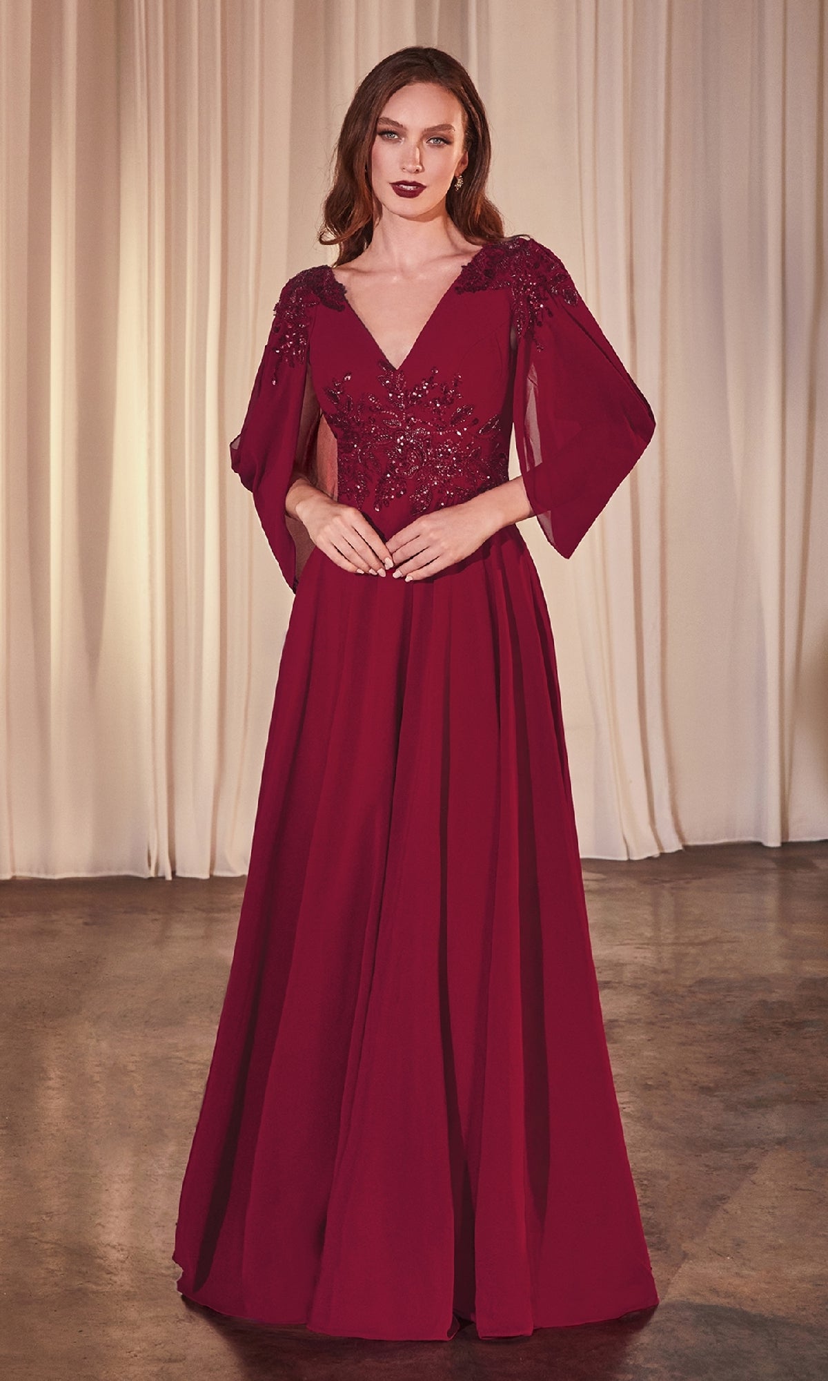 A woman stands on a polished floor in the Ladivine CDS508 Cowl-Sleeve Long Formal Dress, deep red with sheer sleeves and floral accents, gracefully posed before cream-colored curtains.